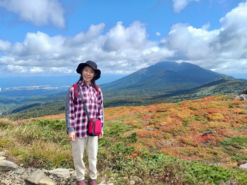 紅葉した山の斜面と遠くの山並みを背に立つ店主